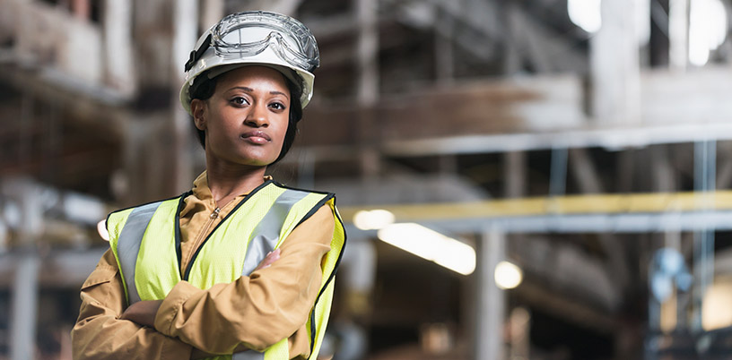 a proud tradeswoman, arms crossed, inside a metal workshop, wearing a helmet and reflective vest